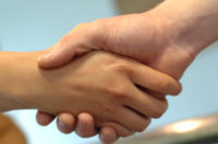 Close-up of two people clasping hands in a welcoming handshake against a soft blue background.