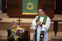 Pastor Chris Ramstad smiles in a white alb and green stole beside an altar bouquet, green paraments behind him, at Immanuel Lutheran Church, Joplin, Missouri.