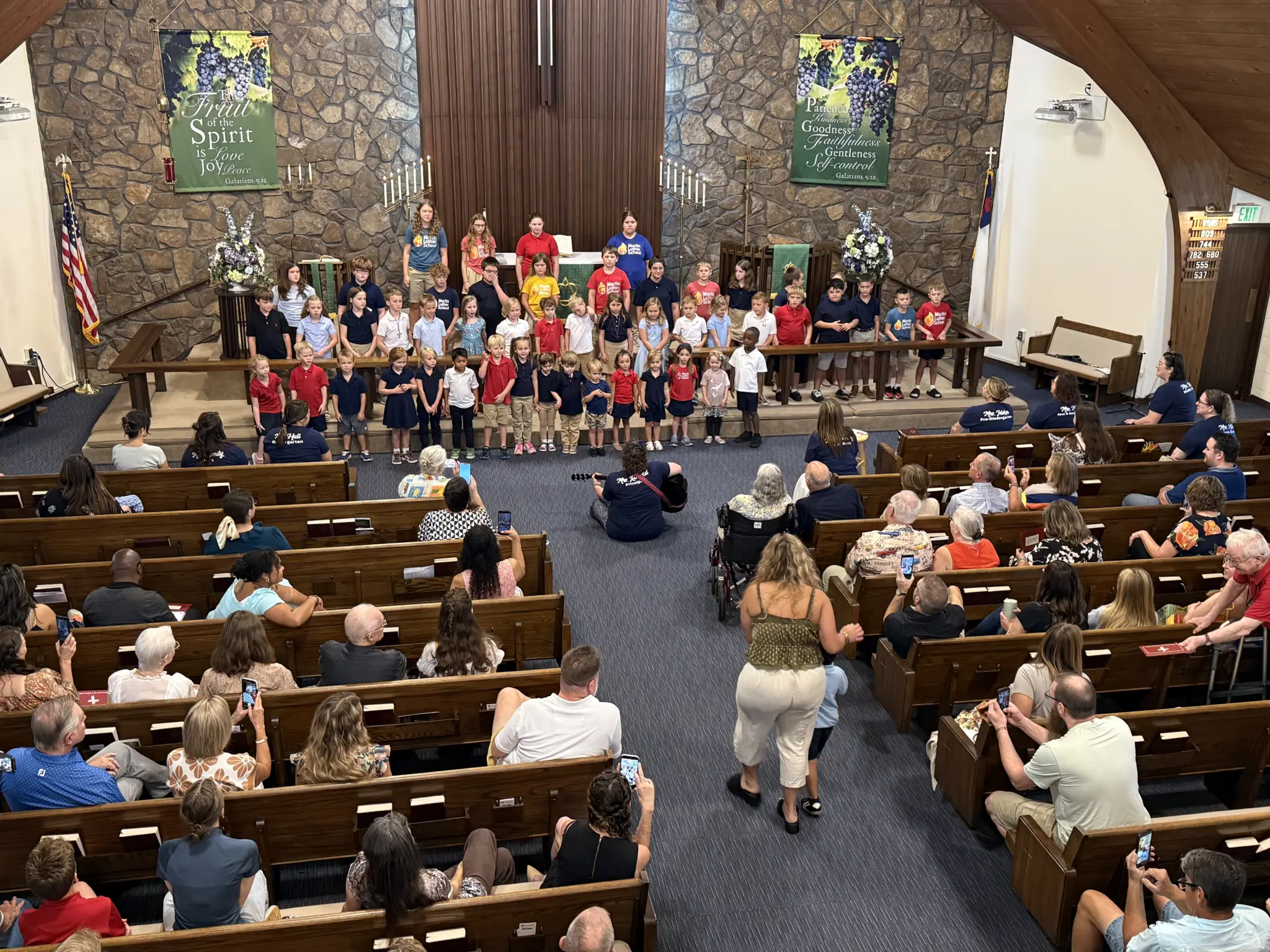 Seventeenth Sunday after Pentecost — September 20, 2026 2 Seventeenth Sunday after Pentecost — Martin Luther School students standing in rows across the chancel steps to sing at the 10:30 service, seen from the balcony, with their teachers behind them, the congregation filling the pews below, and Fruit of the Spirit banners on the stone chancel wall